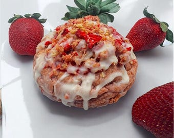 May include: A strawberry-flavored pastry with white icing and strawberry crumble topping, surrounded by fresh strawberries and a green leafy garnish. The dessert is presented on a white plate.