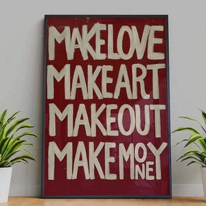 May include: Framed art print with a red background and white text. The text reads "MAKE LOVE, MAKE ART, MAKE OUT, MAKE MONEY". The artwork is in a black frame and is displayed on a wooden surface with plants on either side.