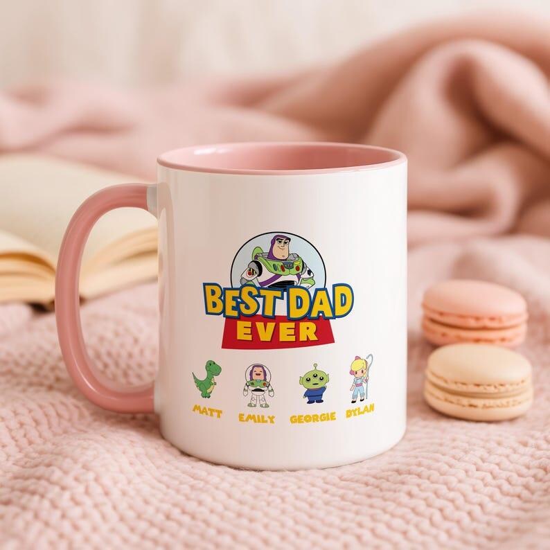May include: A white ceramic mug with a pink interior and handle, featuring a Toy Story graphic with the text BEST DAD EVER. Below the text are four cartoon characters with the names Matt, Emily, Georgie, and Dylan printed underneath them.