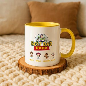 May include: A white ceramic mug with a yellow interior and handle, featuring a graphic of Buzz Lightyear and the text BEST DAD EVER. Below are four small Toy Story character illustrations with the names Matt, Emily, Georgie, and Dylan printed below.