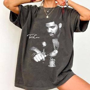 以下が含まれることがあります： 黒と白の男性の画像と「Take Care」のテキストが特徴の濃い灰色のTシャツ。グラフィックは、男性がグラスと台座の上の鳥を持っている様子を示しています。シャツは半袖で、ゆったりとしたフィット感です。