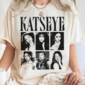 May include: Cream-colored t-shirt featuring the word "KATSEYE" in bold, black letters above six black and white portraits. The portraits are arranged in two rows of three. The shirt has short sleeves and a crew neck.