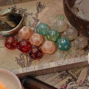 May include: A collection of twelve multi-sided polyhedral dice in various translucent colors including red, peach, and green. Each die features engraved white astrological symbols and numbers, with some faces displaying zodiac signs and others showing numerals.