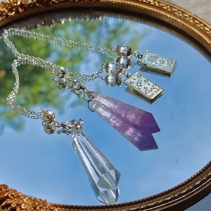 Pode incluir: Um colar de corrente prateada com dois pingentes: uma ponta de cristal transparente facetada e uma ponta de cristal roxa facetada. Inclui dois pequenos berloques retangulares que representam uma mão e estrelas com o texto The Star.