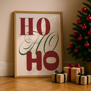Peut inclure: Une affiche encadrée avec les mots "HO HO HO" en rouge et vert, appuyée contre un mur. Un sapin de Noël avec des boules rouges et des cadeaux emballés avec des nœuds sont au premier plan. Les cadeaux sont emballés dans du papier brun.