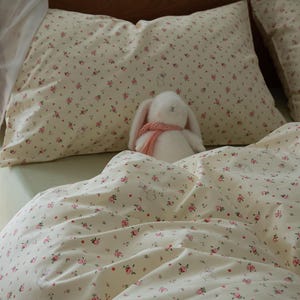 Puede incluir: Juego de cama color crema con estampado floral y de conejos. El juego incluye una funda de almohada, una funda nórdica y un conejito de peluche con una bufanda rosa. El estampado floral presenta pequeñas rosas rosas y hojas verdes.