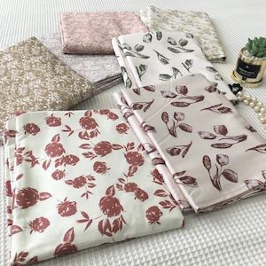 Könnte beinhalten: Eine Sammlung gefalteter Stoffstücke mit floralen Mustern. Die Stoffe weisen verschiedene Designs auf, darunter Rosen, Tulpen und abstrakte Blumendrucke in Braun-, Rosa- und Grüntönen. Die Stoffe sind auf einer weißen Oberfläche angeordnet.