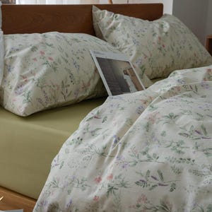 以下が含まれることがあります： 木製のヘッドボードと淡い緑色のフィットシーツを備えたベッド。寝具は、クリーム色の背景にラベンダー、ピンク、緑のアクセントが付いた花柄です。2つの枕と羽毛布団が見えます。