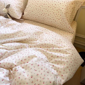Może przedstawiać: Zestaw pościeli w kwiaty. Zestaw zawiera poszwę na kołdrę, poszewki na poduszki i prześcieradło z gumką. Tkanina jest biała z różowymi paskami i małymi różowymi kwiatami. Na łóżku leży pluszowy królik.