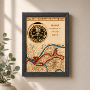 May include: A framed wooden display featuring a Pittsburgh Marathon 2026 medal, a laser-cut map of the city with a red route line and blue river, and text fields for name, grades, and date. The frame is dark gray with a light wood background.