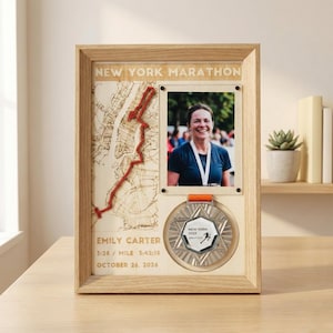 May include: Wooden frame with a map of the New York Marathon route, a photo of a woman, and a medal. The frame includes the text "NEW YORK MARATHON", the runner's name, race time, and date. The medal is silver with an orange ribbon.