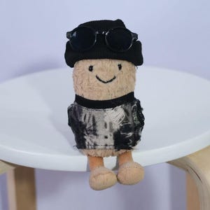 May include: A soft toy figure with a smiling face, wearing a black beanie, sunglasses, and a black and grey jacket. The toy is sitting on a white, round table with light-coloured wooden legs.