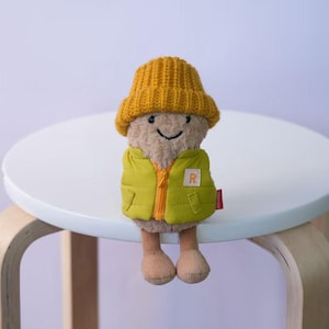Könnte beinhalten: Ein Plüschspielzeug in Form eines Pilzes, das eine gelbe Strickmütze und eine grüne Weste mit einem "R"-Aufnäher trägt. Das Spielzeug hat ein lächelndes Gesicht und sitzt auf einem weißen, runden Tisch.