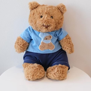 May include: A plush brown teddy bear wearing a light blue t-shirt with a bear applique and navy blue shorts. The stuffed animal is sitting on a white surface against a white background.