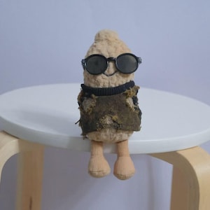 Peut inclure: Une poupée en peluche en forme de cacahuète à la surface beige texturée, portant des lunettes de soleil rondes noires et un gilet marron foncé à l'aspect usé sur un col côtelé noir. La poupée a deux petites jambes beiges aux pieds arrondis.