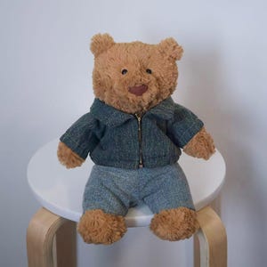 May include: A brown teddy bear wearing a blue tweed jacket with a zip and matching shorts. The bear is sitting on a small white stool with wooden legs. The bear has a brown nose and a stitched mouth.