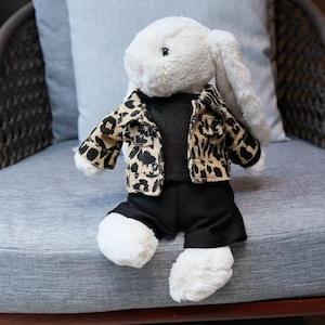 Può includere: Un coniglio di peluche bianco con un top nero a coste, pantaloncini neri e una giacca con stampa leopardata marrone e nera. Il coniglio è seduto su una sedia imbottita grigia.