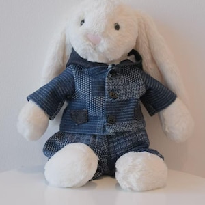 Puede incluir: Un conejo de peluche blanco con una chaqueta azul de retazos y pantalones a juego. El conejo tiene orejas largas, una nariz rosa y ojos negros. El atuendo tiene tres botones y una capucha.