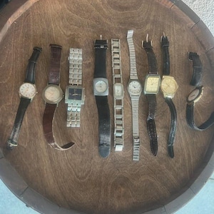 Puede incluir: Una colección de relojes de pulsera vintage con varios estilos de correas, incluyendo cuero y metal. Las esferas de los relojes son redondas y cuadradas, con tonos dorados y plateados. Los relojes están dispuestos sobre una superficie de madera.