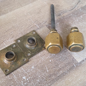 Peut inclure: Ensemble de quincaillerie en laiton antique, comprenant deux plaques carrées avec des ouvertures circulaires et deux boutons octogonaux. L'un des boutons a une longue tige en métal foncé. Les pièces sont patinées.
