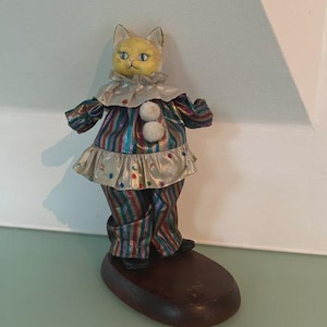 May include: A decorative cat figurine in a clown outfit. The cat has a yellow face and blue eyes, wearing a striped costume with a silver collar and skirt. It stands on a dark brown oval base, a unique decorative item.