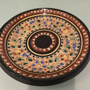 May include: A round, decorative wooden plate with a black base and a detailed, colorful design. The plate features a central black circle, surrounded by intricate patterns in shades of tan, brown, green, blue, and yellow.