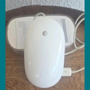 May include: A glossy white wired computer mouse with a centered circular button and a subtle Apple logo embossed on the bottom half. The device features a white USB cable and is positioned in front of its original clear plastic packaging and documentation.