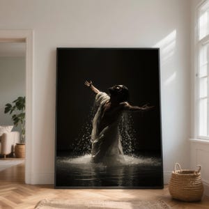 May include: A framed art print depicts a person in a white garment, arms outstretched, standing in water with a dark background. Water splashes around the person, creating a dramatic effect. The artwork is displayed in a bright room with a basket.