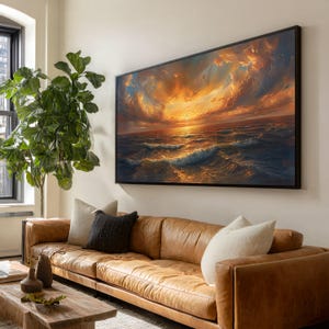 Op de afbeelding: Een groot ingelijst schilderij toont een zeegezicht met een dramatische zonsondergang. De lucht is gevuld met wervelende oranje en gele wolken, die weerspiegelen in de oceaangolven. Onder het kunstwerk staat een bruine leren bank met kussens.
