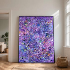 May include: Abstract expressionist painting with a black frame. The artwork features a vibrant mix of colors, including purple, blue, white, yellow, and black, applied with a splatter technique. The painting is displayed on a wooden floor.