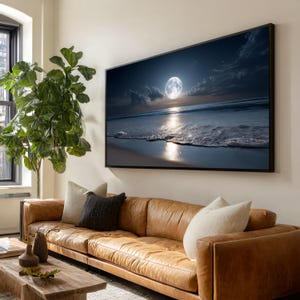 May include: Large framed artwork depicting a nighttime beach scene with a full moon over the ocean. The artwork is displayed above a brown leather sofa with decorative pillows. A large green plant is to the left of the artwork.