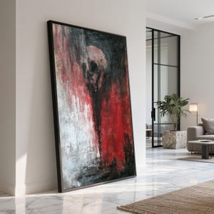 May include: Abstract artwork print with a skull motif in red, black, and white. The painting is framed in a black rectangular frame and leans against a white wall. The artwork has a textured finish.