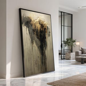 Puede incluir: Impresión artística abstracta enmarcada en negro. La obra presenta una mezcla de tonos negros, dorados y beige con un efecto de pintura texturizada y goteada. La pieza, de gran formato, se apoya en una pared blanca en un espacio de vida moderno.