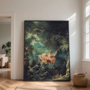 May include: A framed print of a Rococo painting showing a woman on a swing in a verdant forest. The woman is dressed in a pink gown and hat. Other figures are in the scene, and the artwork has a dark frame. The print is displayed on a wooden floor.