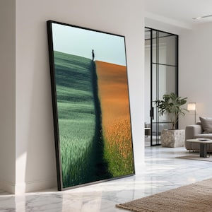 Könnte beinhalten: Gerahmter Kunstdruck, der eine Person auf einem Grat zwischen einem grünen und einem orangefarbenen Feld zeigt. Das Kunstwerk hat einen schwarzen Rahmen und lehnt an einer weißen Wand in einem modernen Interieur.
