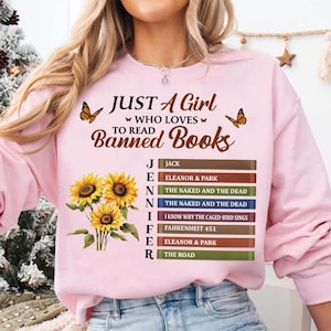 Può includere: Una felpa girocollo rosa chiaro con la scritta "JUST A GIRL WHO LOVES TO READ BANNED BOOKS" in marrone. Sotto, il nome "JENNIFER" è scritto verticalmente accanto a una pila di dorsi di libri colorati, decorata con girasoli gialli.