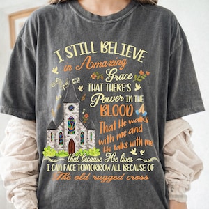 May include: A charcoal gray short-sleeve t-shirt featuring a graphic of a church with stained glass windows. The text reads: I STILL BELIEVE in Amazing Grace THAT THERE'S Power IN THE BLOOD That He walks with me and He talks with me because He lives I CAN FACE TOMORROW ALL BECAUSE OF The old rugged cross.