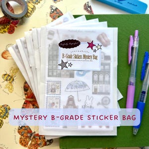 Op de afbeelding: Een stapel doorzichtige enveloppen met stickervellen, voorzien van een witte sticker met de tekst Perfectly Imperfect! journabelle B-Grade Stickers Mystery Bag. De vellen bevatten illustraties van gebouwen, paraplu's en konijnen.