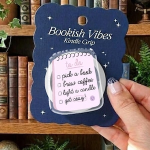 Puede incluir: Un agarre para Kindle azul marino con el texto "Bookish Vibes" y una lista de tareas: elegir un libro, preparar café, encender una vela y ponerse cómodo. El agarre se sostiene frente a una estantería llena de libros.