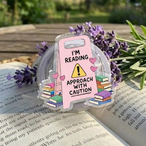 May include: A pink acrylic sign with the text "I'M READING APPROACH WITH CAUTION" and heart and book graphics. The sign is placed on an open book with lavender sprigs in the background. The sign is approximately 3 inches wide.
