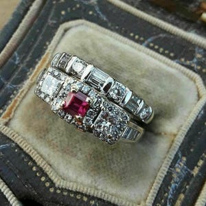 Pode incluir: Dois anéis ornamentados com diamantes e um rubi central são exibidos em uma caixa de joias vintage. Os anéis apresentam uma mistura de diamantes quadrados e baguette, cravejados em uma banda de prata ou ouro branco. A caixa é escura com um fecho dourado.