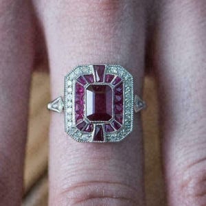 Peut inclure: Bague en argent ornée d'un rubis rectangulaire central, entouré de petits rubis et de pierres claires. La bague présente un design rectangulaire orné. La bague est portée sur un doigt.