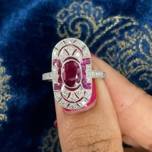 Peut inclure: Une bague ovale allongée présentant un rubis rouge profond de taille ovale au centre, entouré d'une monture géométrique argentée. Le cadre est orné de petits rubis carrés et de nombreuses minuscules pierres blanches disposées symétriquement.