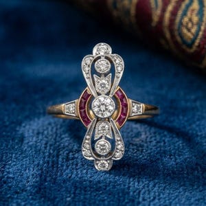 Può includere: Un anello in oro con un elemento centrale verticale allungato incastonato con molteplici diamanti rotondi taglio brillante e due rubini curvi taglio baguette. La struttura metallica presenta filigrana argentata e bordi milgrain.