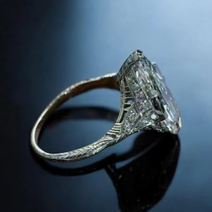 Op de afbeelding: Een ring in antieke stijl met een grote, rechthoekige, heldere edelsteen. De band en zetting van de ring zijn ingewikkeld gedetailleerd met kleinere stenen en decoratieve patronen. De ring staat tegen een donkere, reflecterende achtergrond.