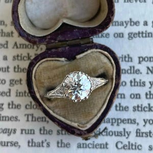 May include: A sparkling diamond ring with a large, round center stone is displayed in a heart-shaped, purple velvet box. The ring has intricate detailing on the band and side stones, showcasing a classic, elegant design.