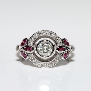 May include: A silver ring with a central, round, clear gemstone encircled by a halo of smaller clear stones. The ring features ruby-colored accents on each side, set in a floral design. The ring is displayed on a white surface.
