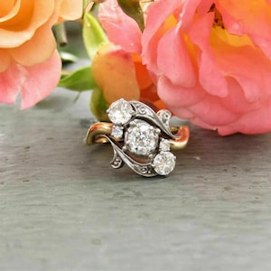 Könnte beinhalten: Ein Ring im Vintage-Stil mit einem goldenen Band und silbernen Filigranarbeiten. Der Ring hat einen großen, runden, klaren Edelstein in der Mitte, umgeben von kleineren Edelsteinen. Der Ring ist vor einem Hintergrund aus rosa und orangefarbenen Rosen platziert.