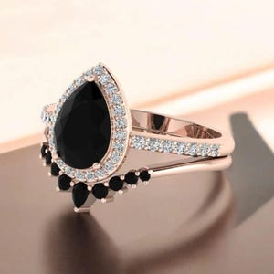 May include: Rose gold ring set with a pear-shaped black gemstone, encircled by small white stones. The band is embellished with more white stones. A second band with black stones completes the set.