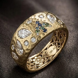 May include: A wide gold band ring with a hammered texture, featuring inlaid clear pear and round-cut stones, a small insect motif with blue and green stones, and an interior engraving that reads 18K AU with floral filigree detailing.